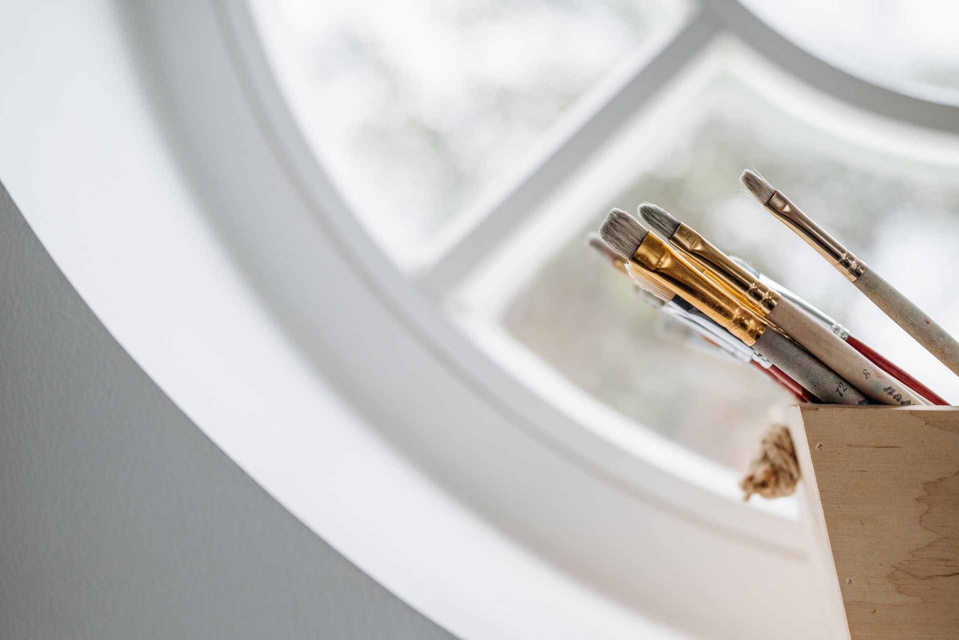 A collection of paintbrushes in a wooden box near a large circular window offering soft natural lighting.