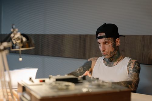 Free Tattooed man in a white tank top and black cap focuses intensely at a desk indoors. Stock Photo