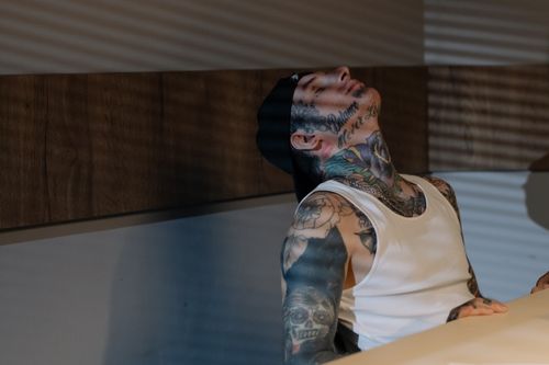 Free A heavily tattooed man in a tank top sits back indoors, relaxed and contemplative. Stock Photo