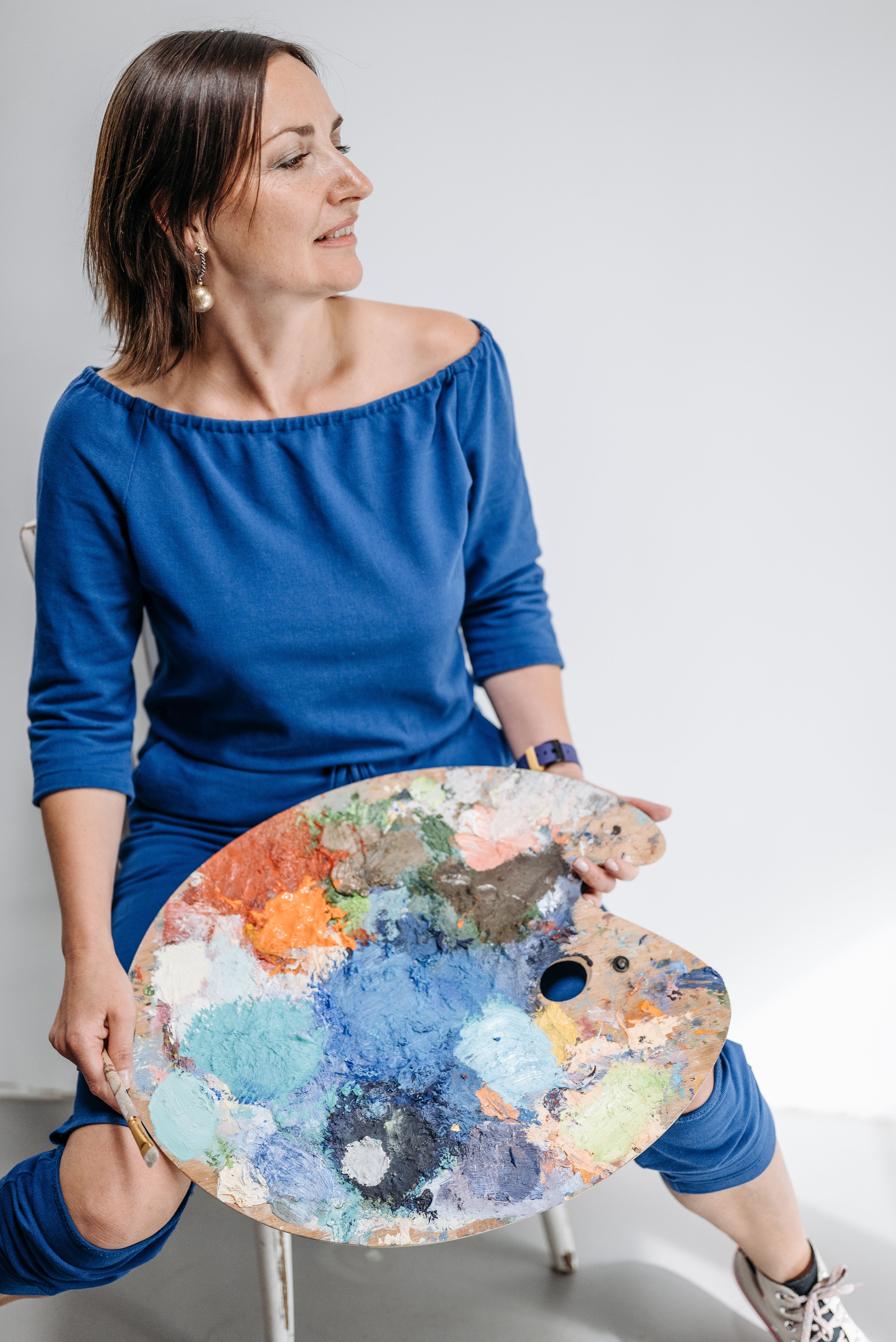 Female artist in blue holding a vibrant paint palette in an art studio.