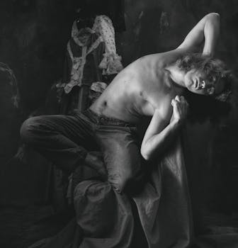 A shirtless man artistically posing in a monochrome setting with vintage attire in the background.