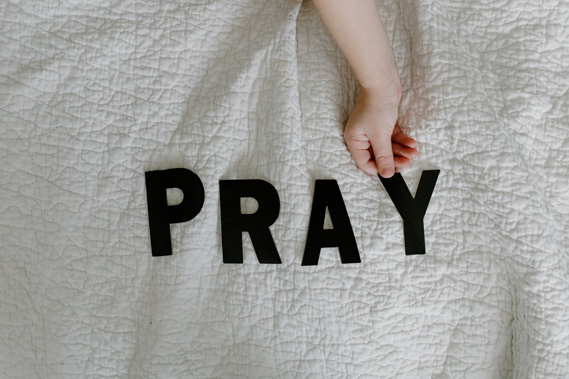 A child's hand placing black letters spelling 'PRAY' on a white quilted blanket.