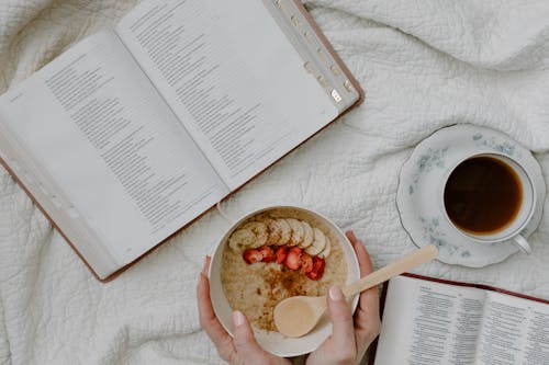 A Person Holding a Bowl of Cereal Beside the Open Book and Coffee