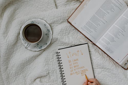 A Person Writing on the Notebook Beside a Book and Coffee