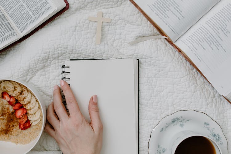 A Person Holding A Blank Notebook Beside The Books And Bowl Of Cereal