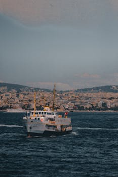 A vibrant ferry boat navigating a deep blue sea with a bustling cityscape in the background.