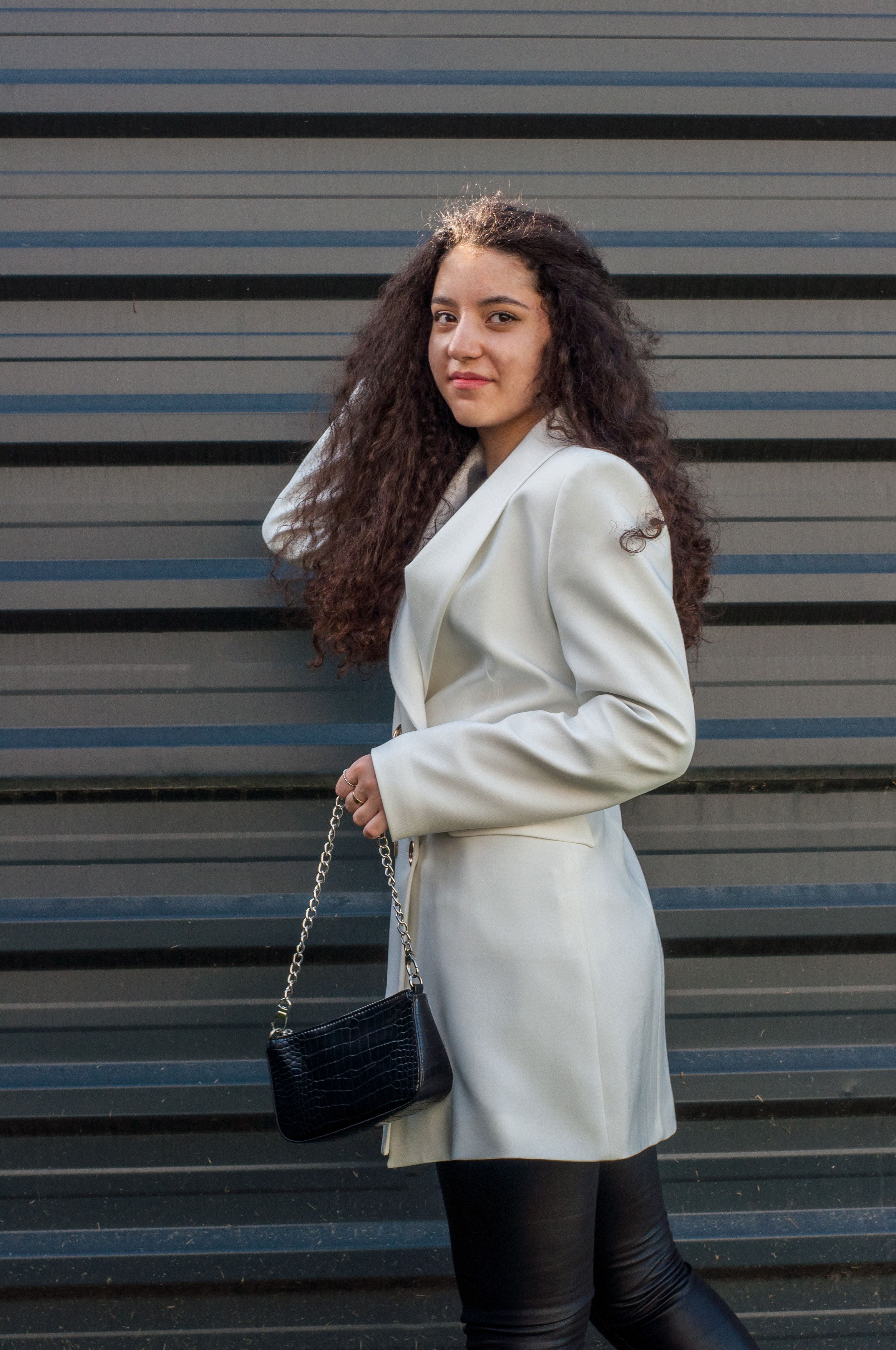 Uma mulher com cabelo cacheado se destaca de forma estilosa contra uma parede texturizada, segurando uma bolsa preta moderna.