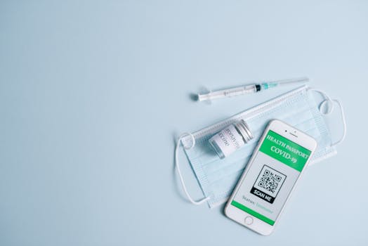 Top view of Covid-19 health passport on smartphone with vaccine, syringe, and mask on blue background.