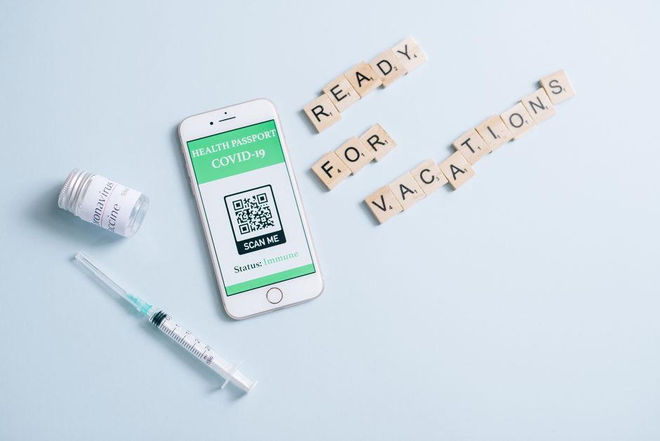 Smartphone displaying COVID-19 health passport with scrabble tiles spelling 'Ready for Vacations', vaccine vial, and syringe.