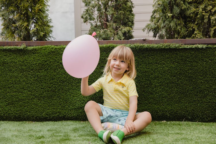 A Child In Yellow Shirt Sitting On Green Grass Holding Pink Balloon