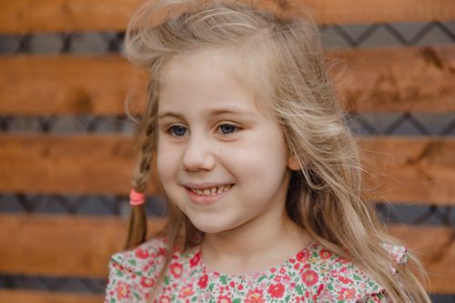 Free Candid portrait of a smiling girl with blonde hair and floral dress against a wooden background. Stock Photo