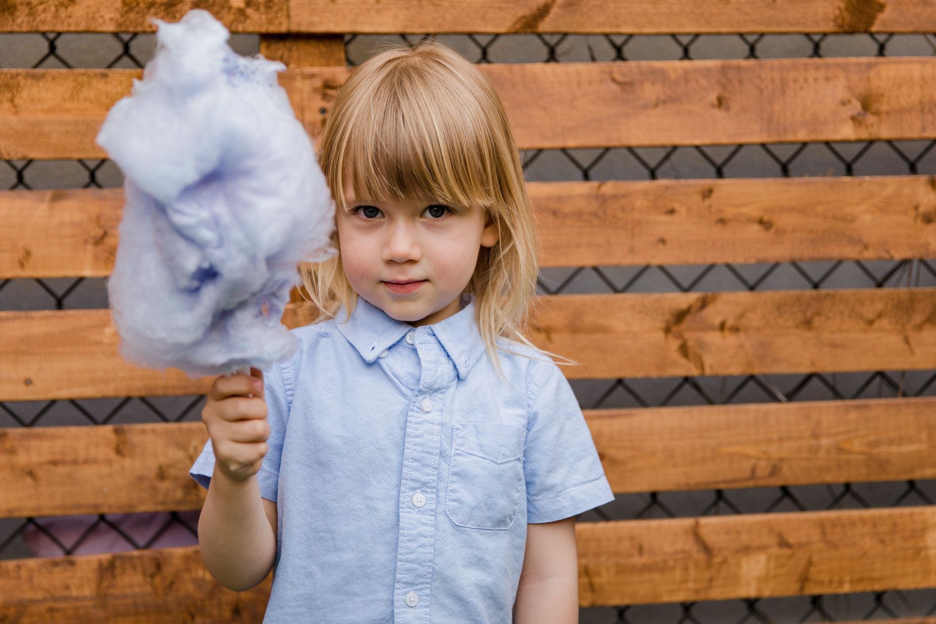 candy floss station events cairo — A young child holding blue cotton candy against a wooden fence, enjoying an outdoor summer