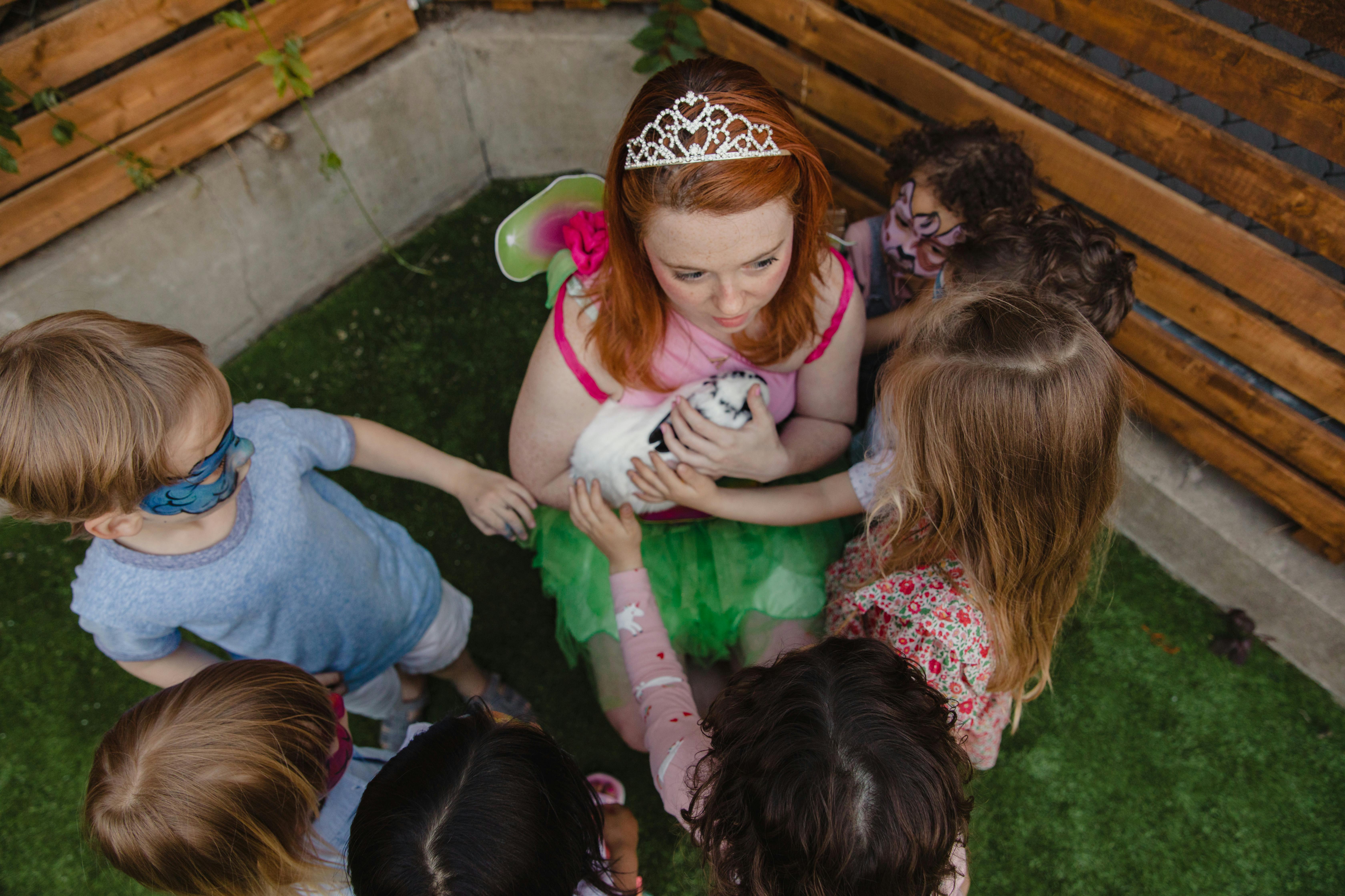 A lively group of children with face paint gather around a woman dressed in costume holding a rabbit at a birthday party.