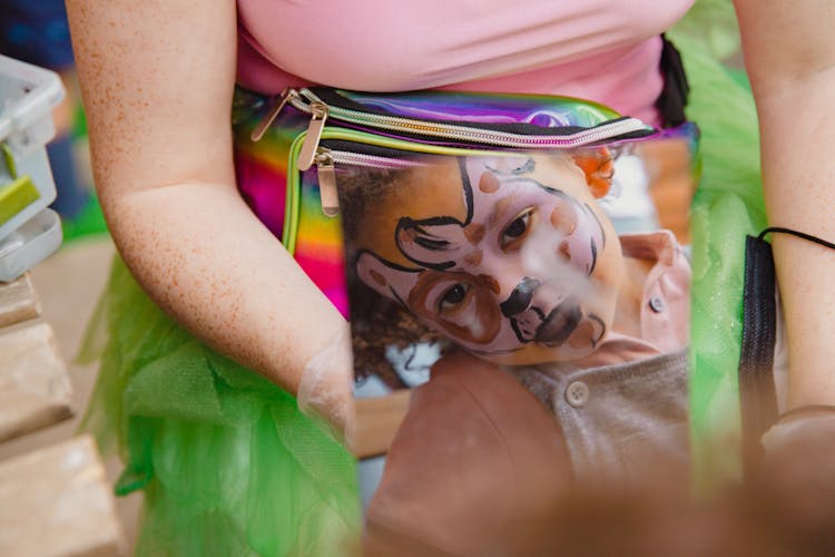 A Kid With Face Paint Looking At A Mirror Near A Person With A Colorful Belt Bag