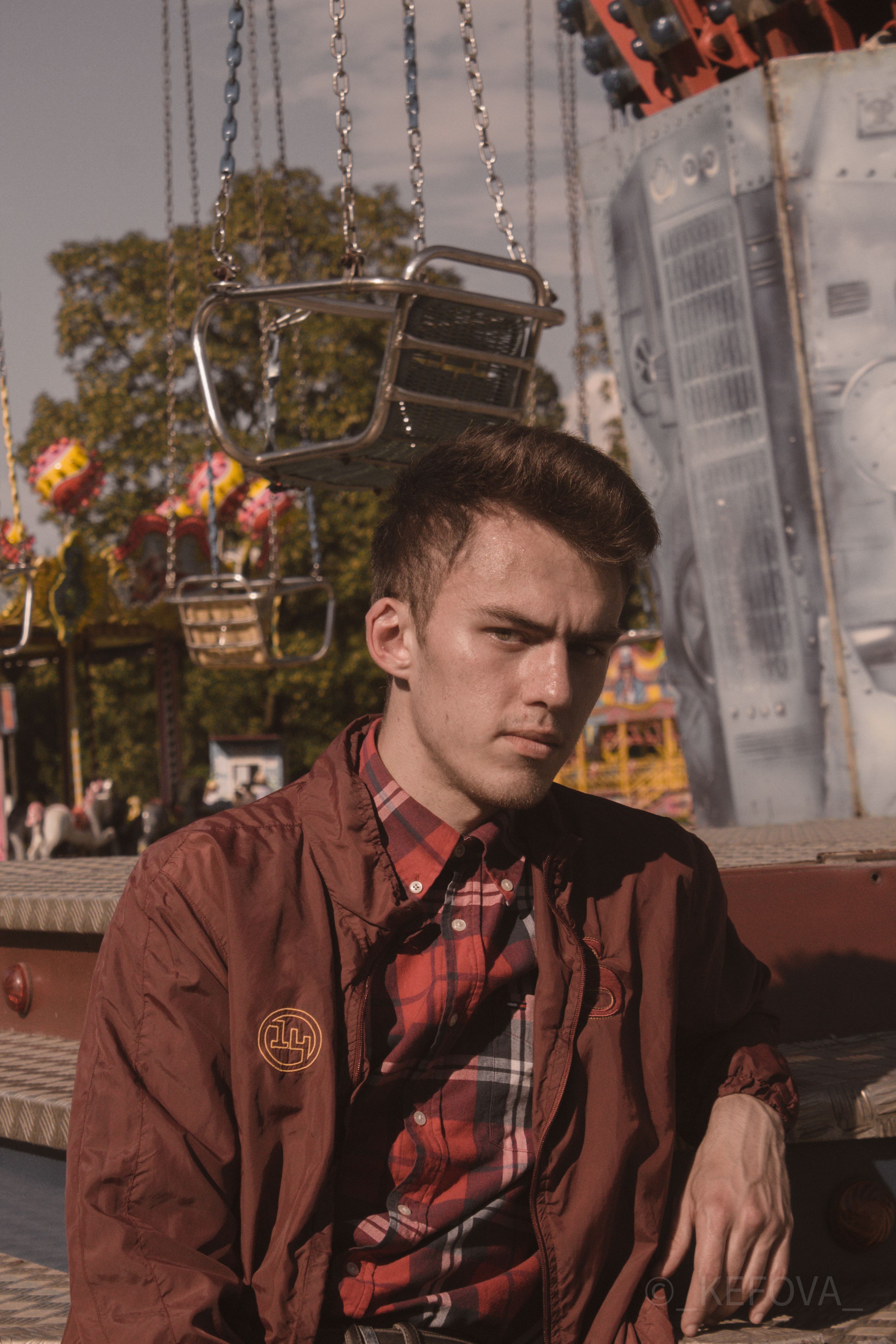 Portrait of a young man in plaid jacket posing at an amusement park with rides.