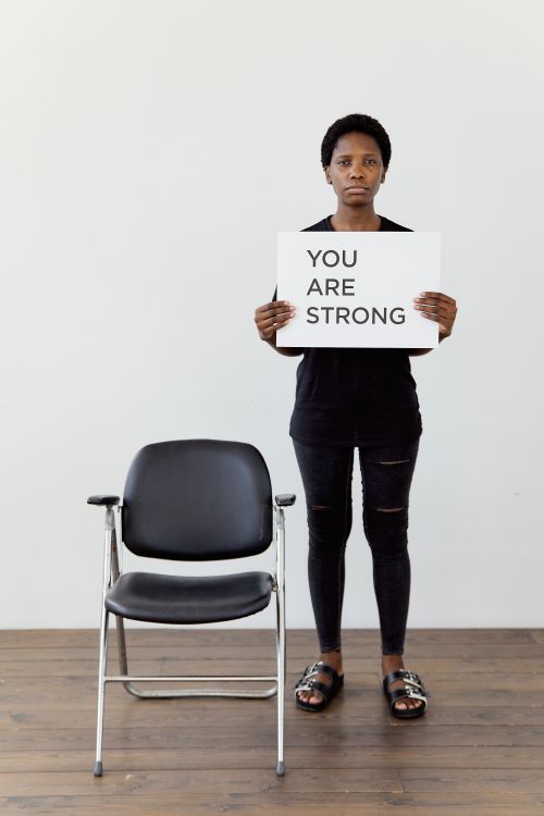 Free African American woman standing indoors, holding a 'You Are Strong' sign, illustrating empowerment. Stock Photo