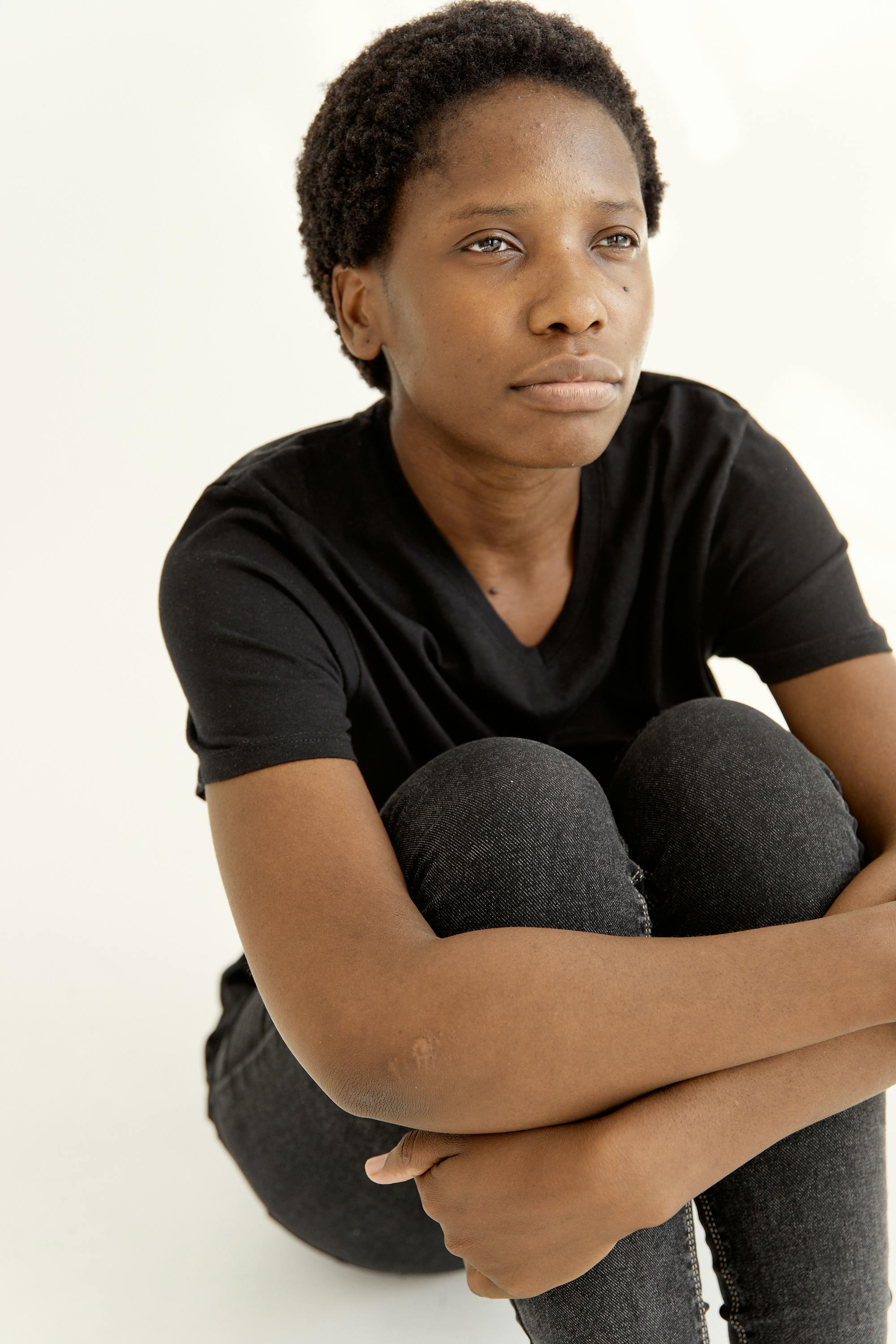 African American woman in black shirt and denim pants sitting on the floor, looking upwards with a contemplative expression.