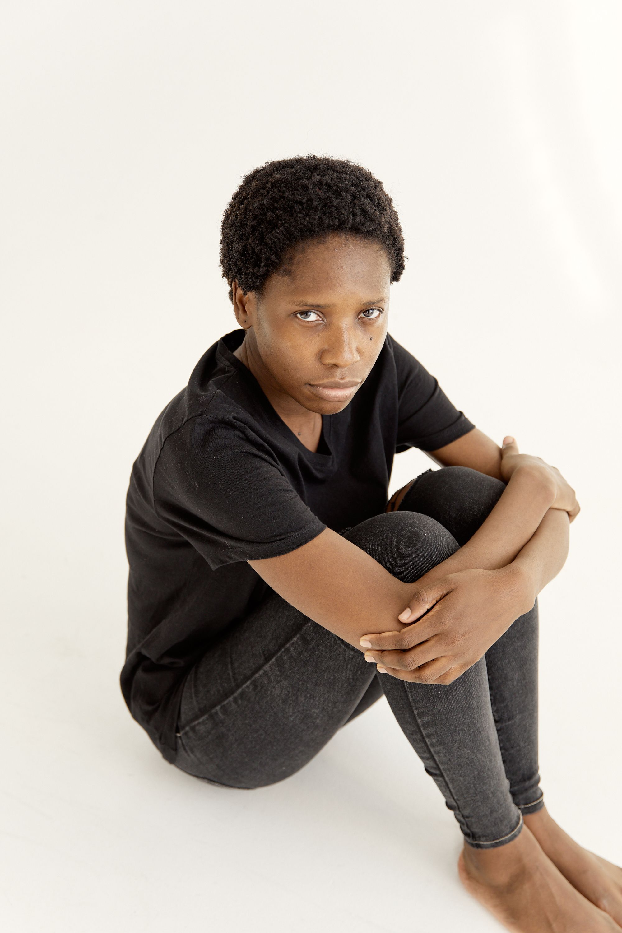 A thoughtful young woman sitting cross-legged on a white surface, looking at camera.