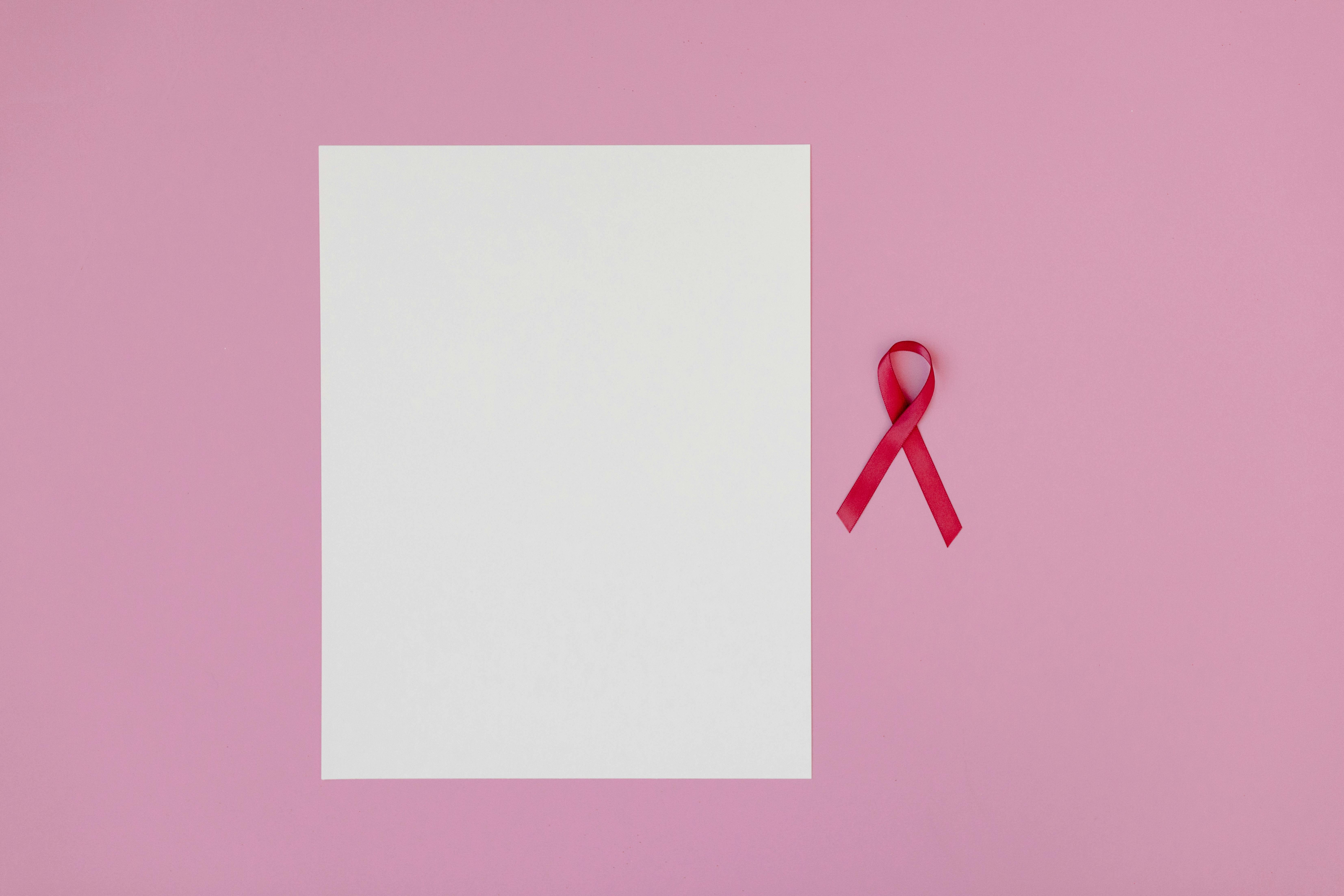 Pink background with a blank paper and a red ribbon symbolizing breast cancer awareness.