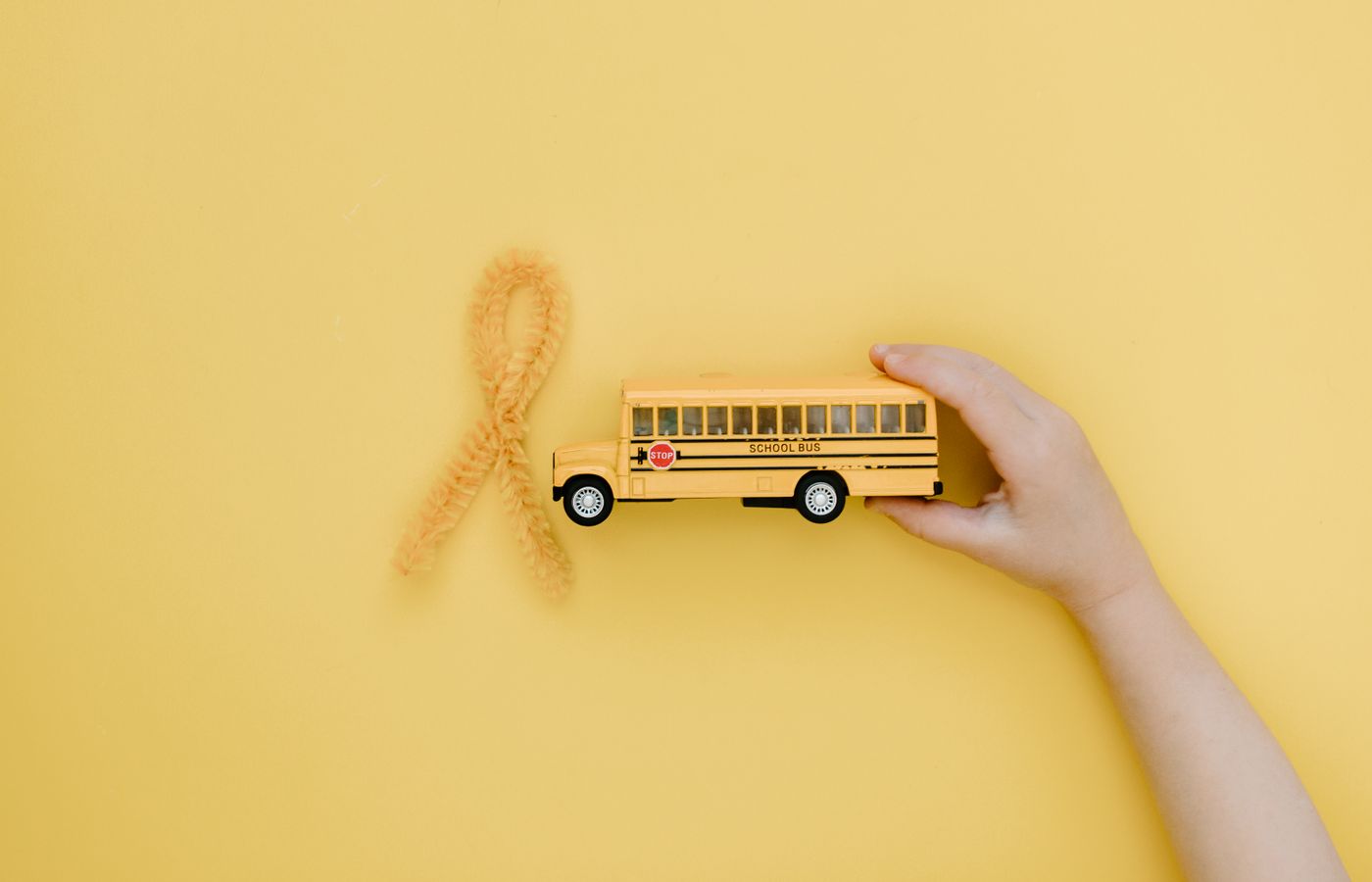 Hand holding toy school bus near yellow ribbon for cancer awareness on yellow background.