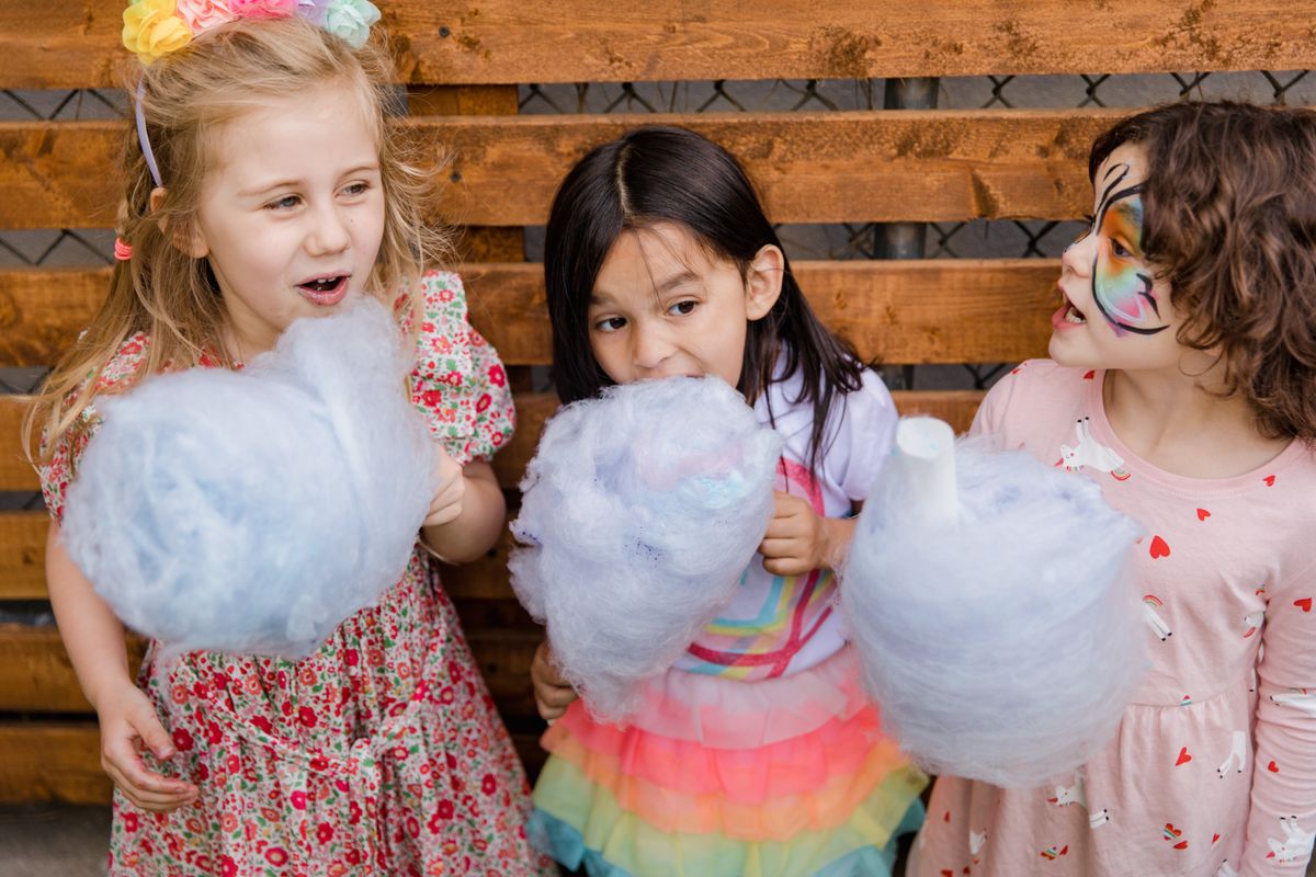 Three children with face paint enjoy cotton candy at an outdoor celebration.