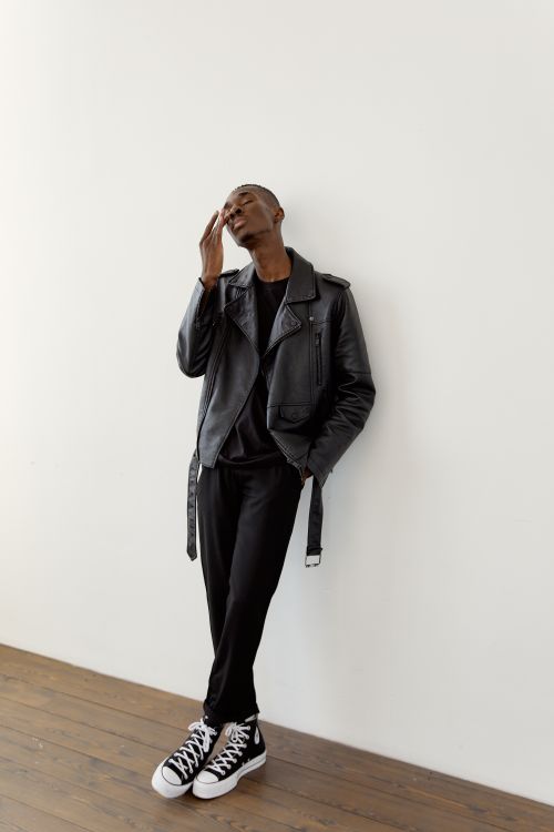 Free A fashionable man leaning against a white wall wearing a leather jacket and sneakers. Stock Photo