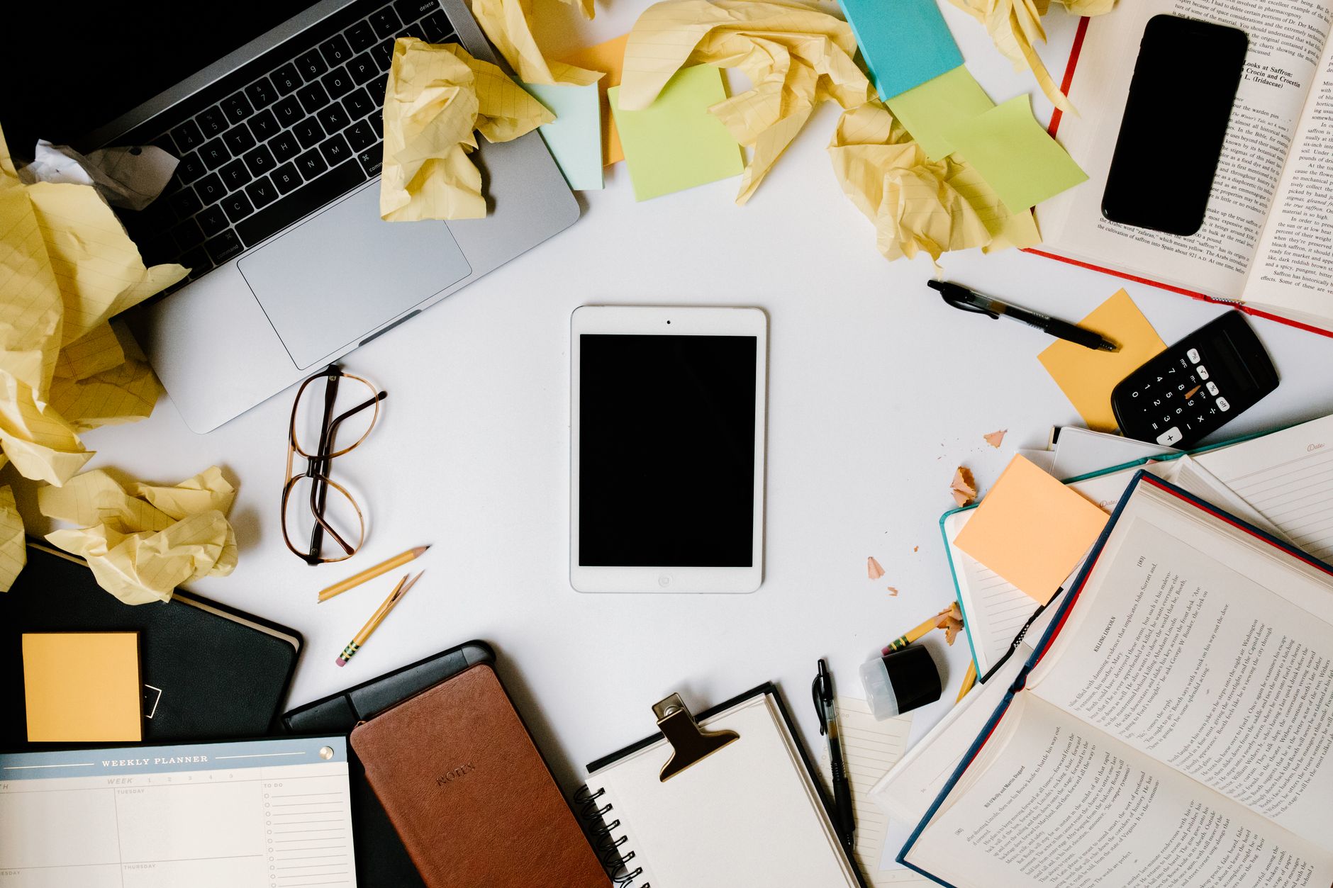 Overhead view of a cluttered workspace with gadgets, crumpled papers, and office supplies.