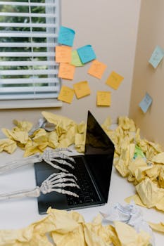 Creative depiction of a skeleton working on a laptop amidst cluttered papers in an office setting.