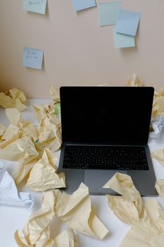 A messy desk with a laptop surrounded by crumpled papers and sticky notes on a wall.