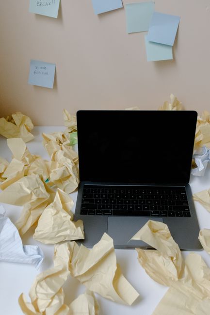 A messy desk with a laptop surrounded by crumpled papers and sticky notes on a wall.