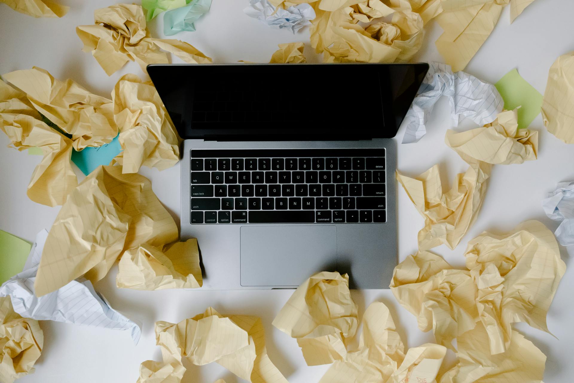 A disorganized workspace with a laptop amid crumpled papers, top view.