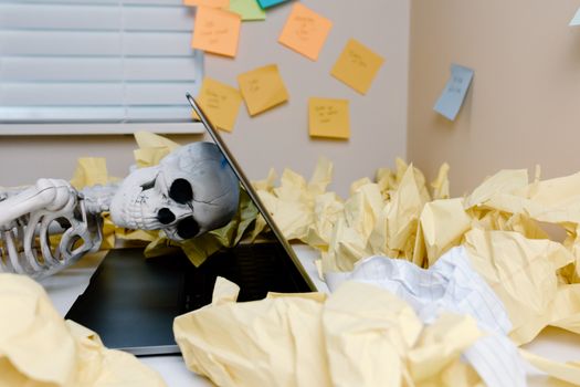 A humorous skeleton slumped over a laptop among crumpled papers in an office setting.