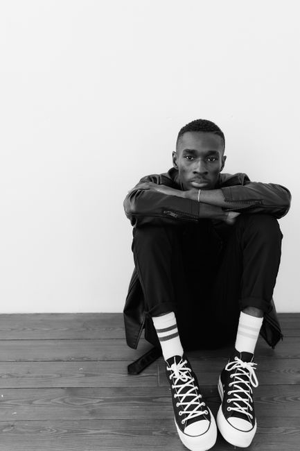 Stylish black and white portrait of a man in a leather jacket sitting on a wooden floor.