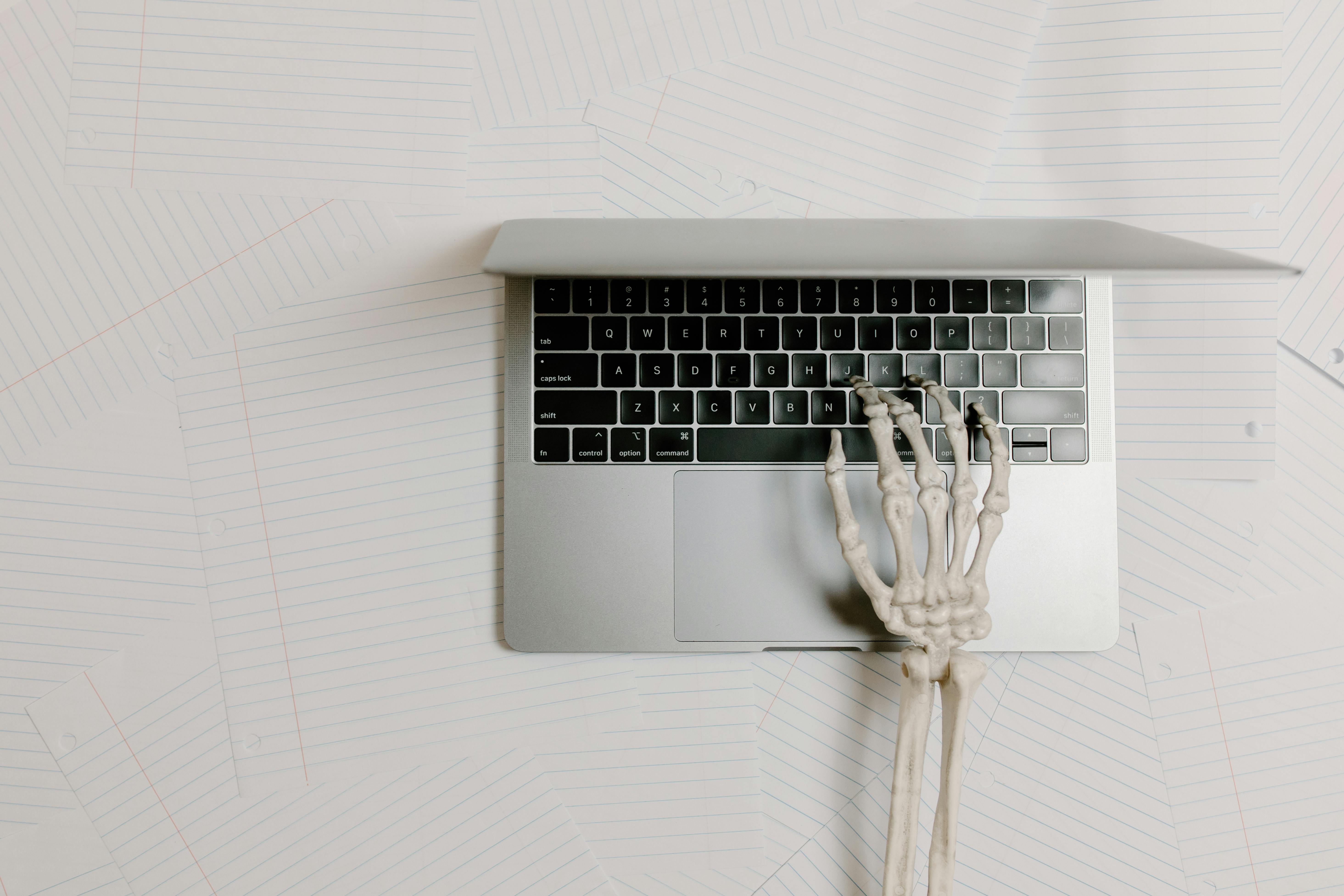 A conceptual overhead view of a skeleton hand typing on a laptop over scattered papers.