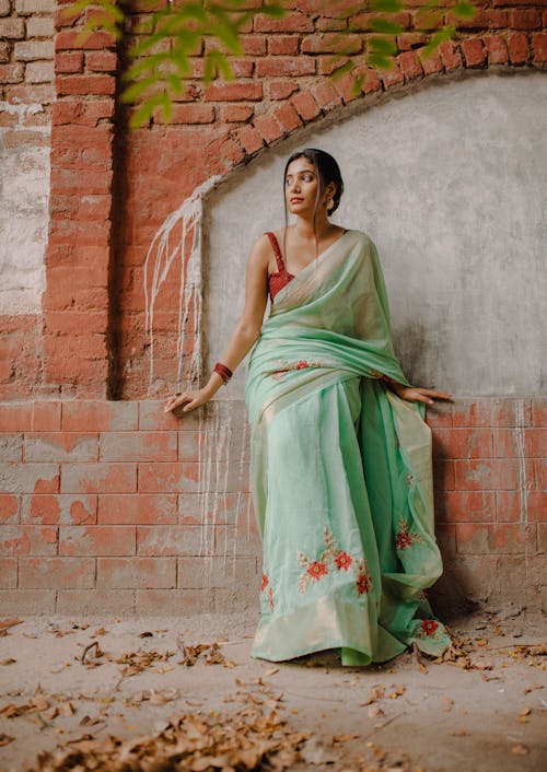 Woman in Traditional Green Floral Dress Leaning on Brown Brick Wall