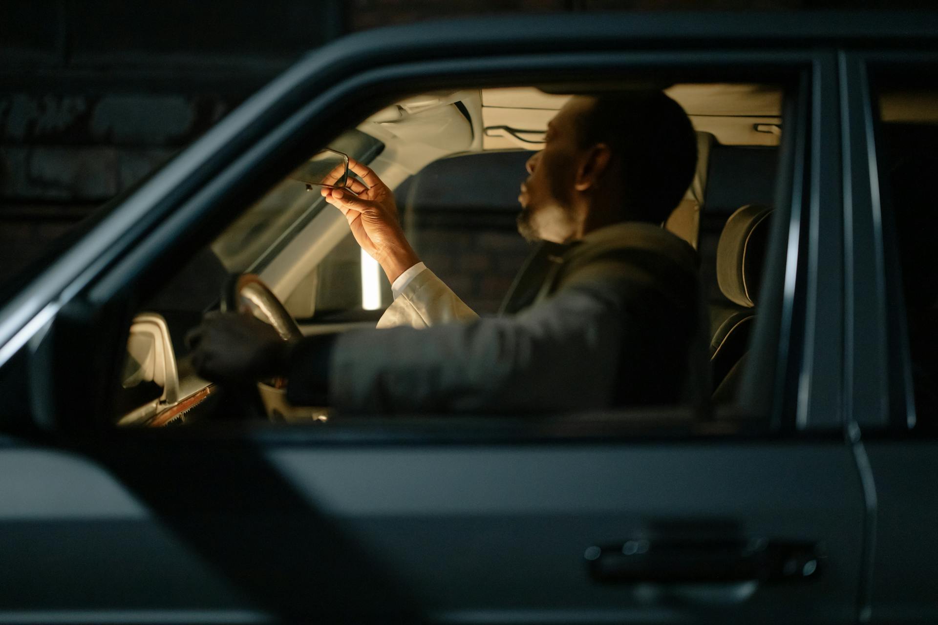 A man sitting inside a car at night adjusting the rearview mirror. Interior illuminated with selective focus.