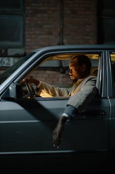 African American man driving silver car at night, arm resting out the window, illuminated by streetlights.