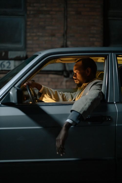Free African American man driving silver car at night, arm resting out the window, illuminated by streetlights. Stock Photo