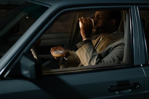 A Man Holding a Donut and Drinking Coffee while Sitting in a Car