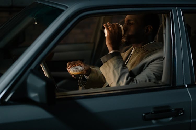 A Man Holding A Donut And Drinking Coffee While Sitting In A Car