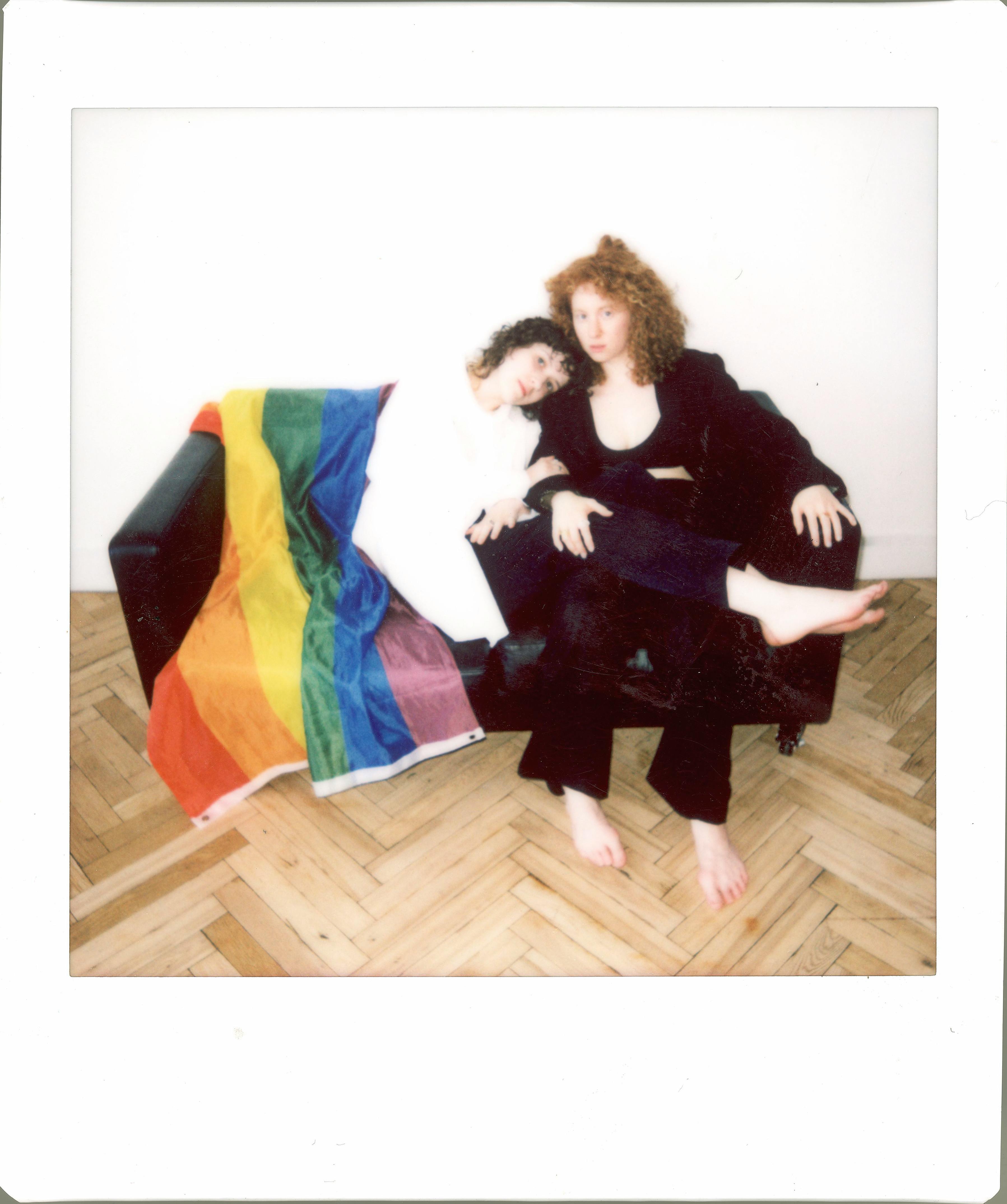 A lesbian couple relaxes on a couch indoors with a rainbow flag, symbolizing LGBT pride.