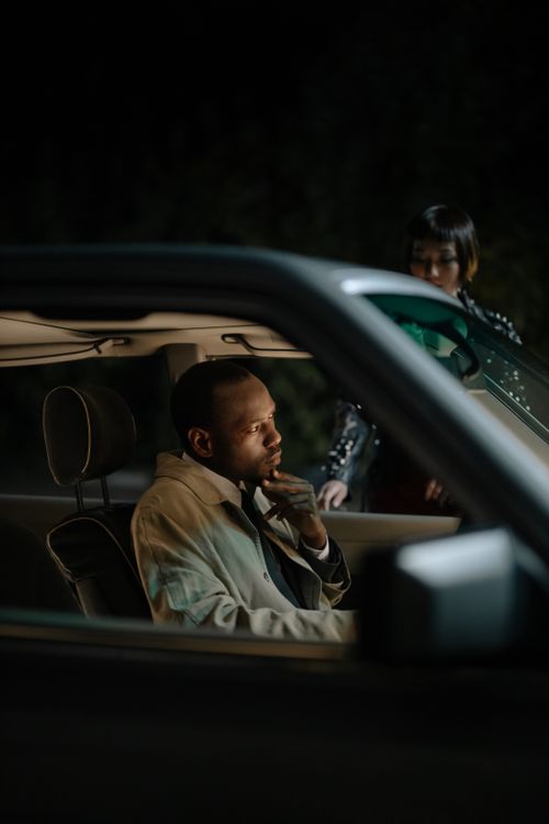 Free Man thoughtfully seated inside a parked car at night, with a person approaching outside the window. Stock Photo