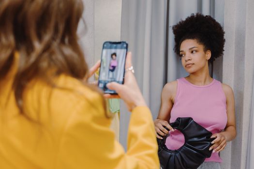 Woman in pink top poses with handbag while another takes a smartphone photo in a changing room.