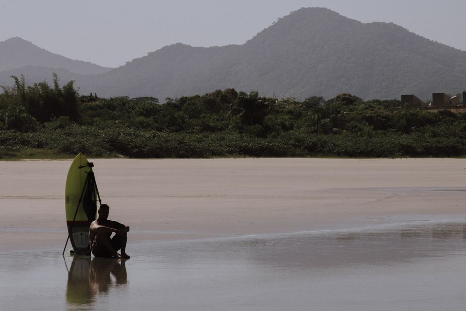 A lone surfer sits by his board, enjoying a peaceful beach with mountains in the backdrop.
