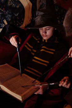 Child in wizard costume studying spells from an open book with a magic wand indoors.