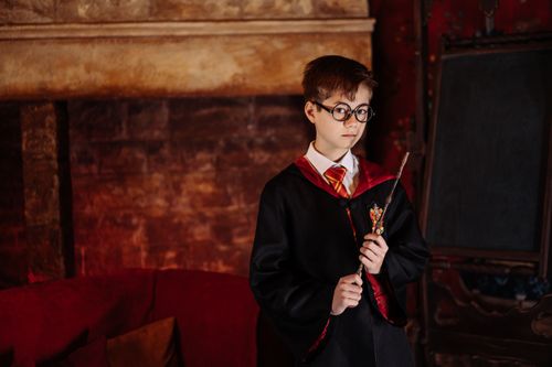 Free Boy dressed as a wizard with eyeglasses and robe holding a wand in front of a fireplace. Stock Photo