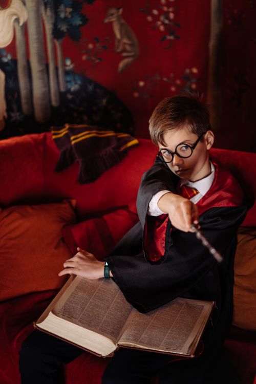 Free Child in wizard robe with glasses, holding wand and book on a cozy sofa. Stock Photo