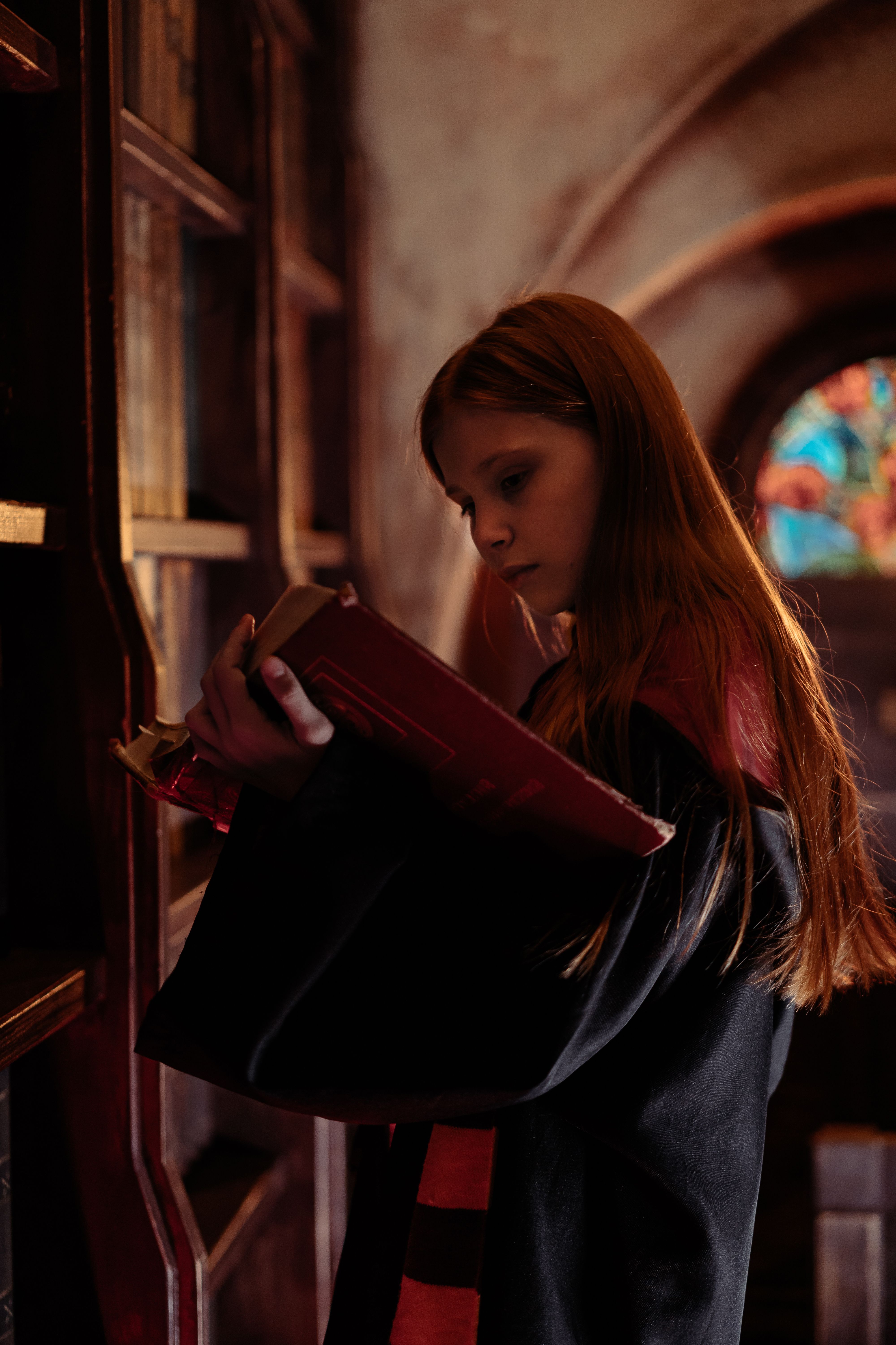 A young girl in a historical setting reading an old book in a warmly lit library.