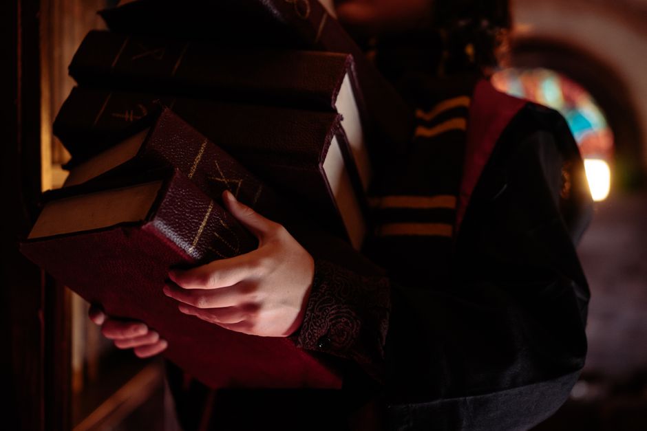 A student in a dark robe carrying a stack of old books in a dimly lit library, evoking mystery.