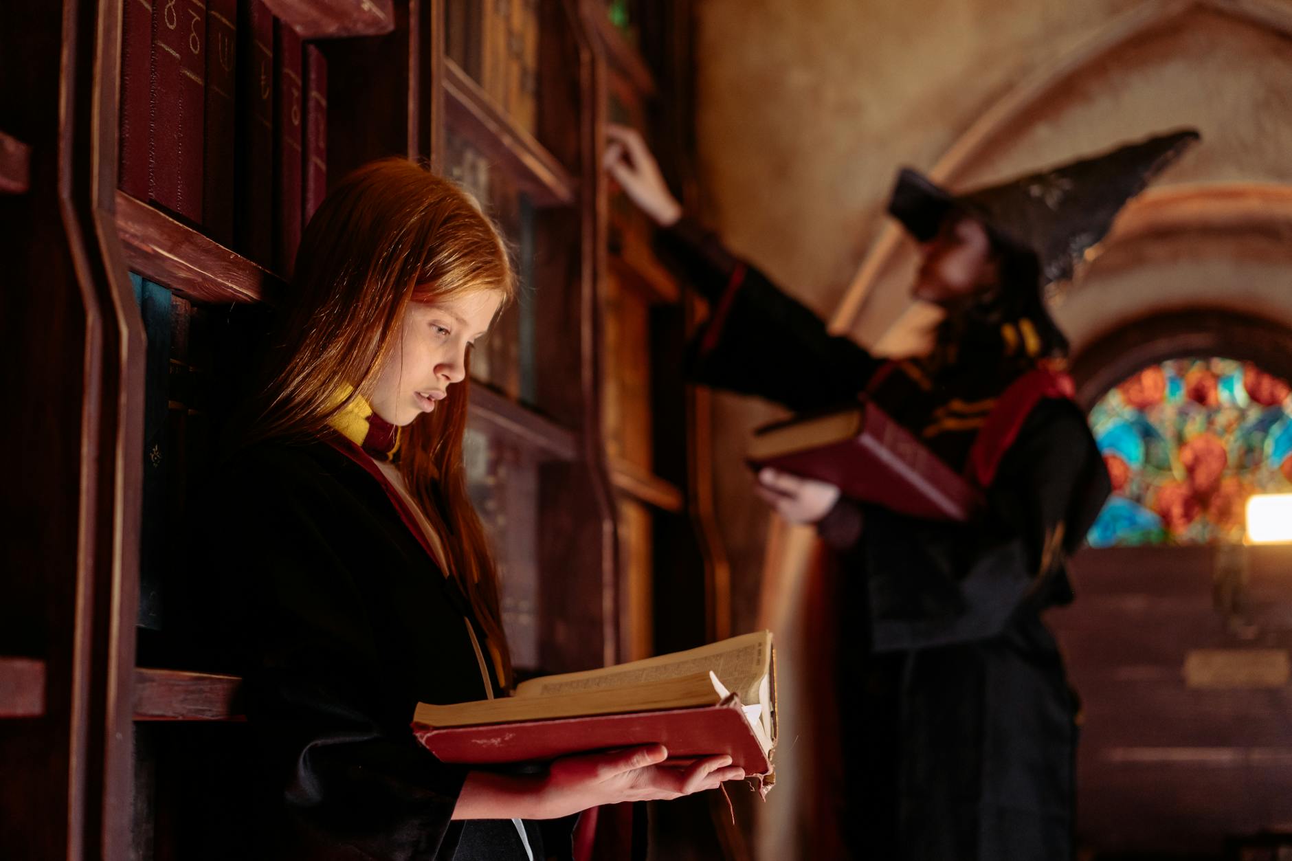 A young girl in a wizard costume reads an ancient book in a magical library setting.